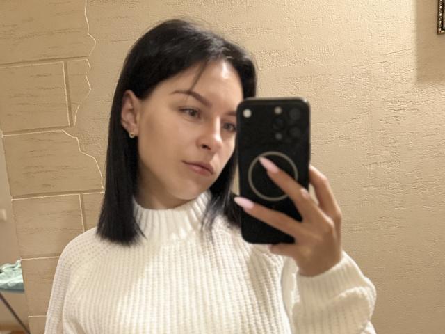 EmmaNoa, com cabelo preto, suéter branco texturizado e unhas rosa, tirando uma selfie no espelho com um celular preto, mostrando seu rosto fofo e pequenos brincos..