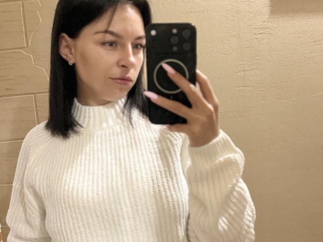 EmmaNoa, con un seno di media grandezza, indossa un maglione bianco a coste, tiene un telefono, sta scattando un selfie allo specchio, ha i capelli neri, la pelle chiara e un sorriso leggero, in un bagno..