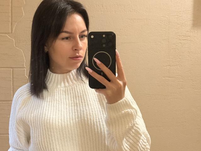 EmmaNoa met recht zwart haar, die een witte gebreide coltrui draagt, een zwarte telefoon met een ring vasthoudt en een spiegel-selfie maakt, heeft een neutrale uitdrukking en een lichte huid..