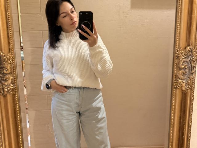EmmaNoa, con la pelle chiara, i capelli scuri, un maglione a coste bianco a collo alto, jeans azzurri a vita alta, in piedi, che si scatta un selfie allo specchio, con una mano in tasca, uno specchio con cornice dorata, un telefono nero..