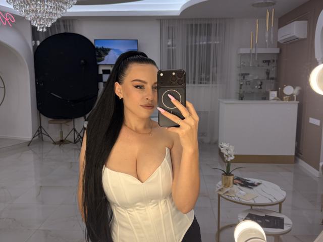 EmmaNoa, met lang zwart haar, draagt een witte top zonder bandjes die haar grote borsten laat zien, en ze maakt een foto van zichzelf in een spiegel met een smartphone in een moderne, goed verlichte kamer. Ze glimlacht en ziet er zelfverzekerd uit..