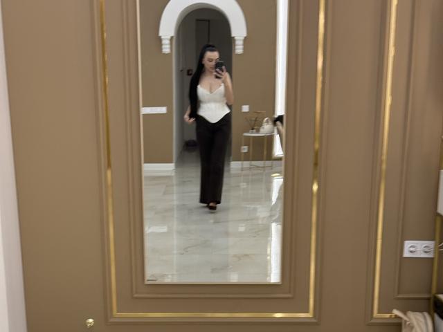 EmmaNoa with dark hair, white bustier, black pants, taking a mirror selfie, showing her tits, holding a phone, standing on shiny marble floor, brown door in the foreground.