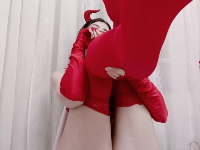 WollyMolly in red devil costume with horns, holding her red-covered butt in front, wearing red gloves, shot from low angle, showing her face partially covered by her hand.