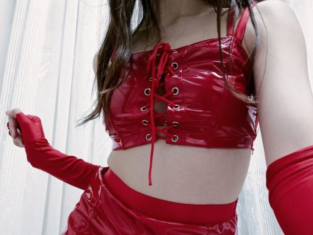 WollyMolly portant un crop top rouge brillant en latex avec laçage sur le devant, un short rouge taille haute en latex, et des gants rouges en latex, laissant apparaître son ventre et sa taille fine..