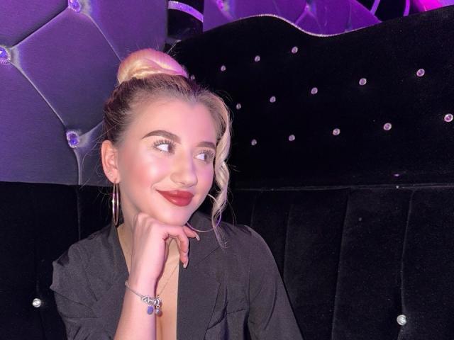 LennieClemens con moño rubio, lápiz labial rojo, camisa negra, aros grandes y pulsera, con la cabeza apoyada en la mano, sonriendo, fondo morado y negro..