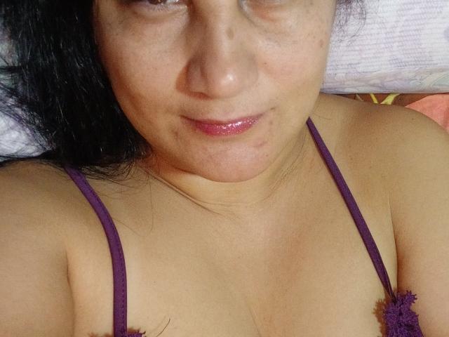 KarinaCecillia with a slightly shiny, pinkish lip, lying down, wearing a purple spaghetti strap top, showing her upper chest and neck, with dark hair on a white pillow.