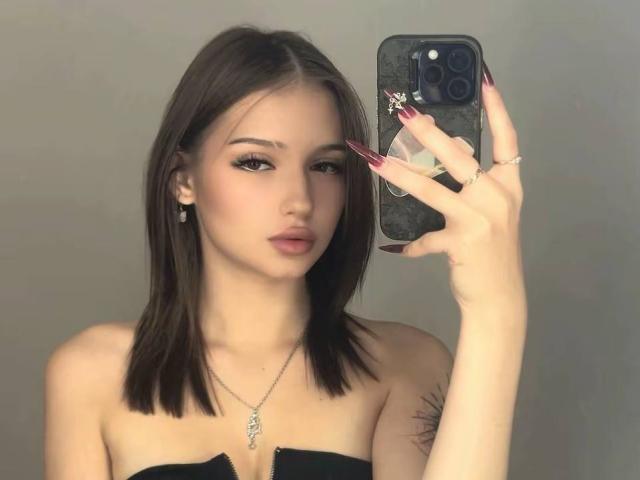 ShanekaKonig with medium-length brown hair, holding a phone with long red nails, wearing a strapless black top, necklace, and earrings, looking at the camera.