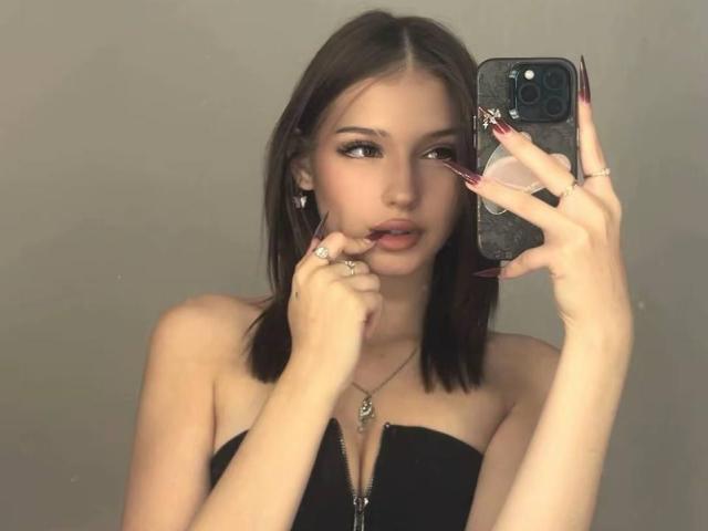 ShanekaKonig with brown hair, wearing a black strapless top, holding a phone with long pink nails, touching her lip with her finger, looking at the camera.
