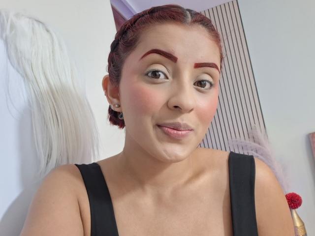 LizBonnet with red braided hair, thick eyebrows, pink cheeks, small nose, and black top, showing a cute smile, has medium-sized tits and a cute face.