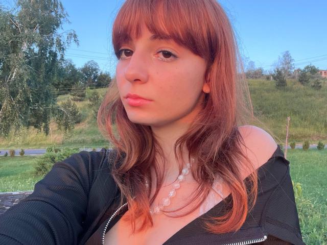 LeomaHaq has red hair, bangs, and a pensive look. She's wearing a black jacket with a low neckline, showing her tits, and a white bead necklace.