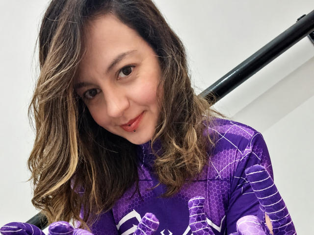 ArianaBlonde with wavy brown hair and a nose piercing, wearing a purple Spider-Man suit, smiling at the camera, holding a black pole in her right hand.