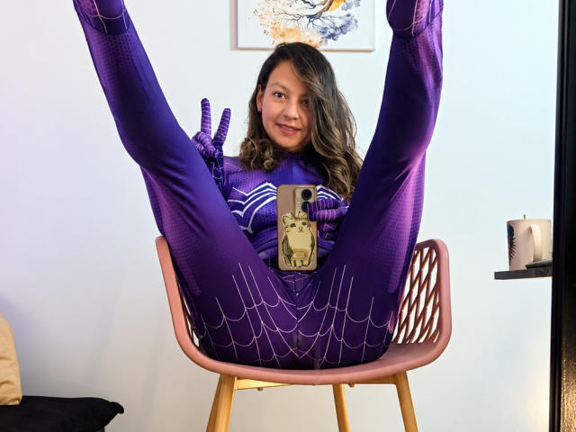 ArianaBlonde in a purple Spider-Man suit, legs spread, holding a phone with a Spider-Man case, sitting on a pink chair, winking at the camera. Her pussy is covered.