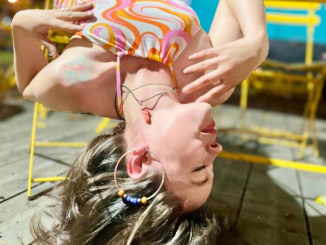 Iniridax, upside down, wearing a colorful, swirly top, with hands near her face, has a tattoo on her arm, hoop earrings, and is on a wooden floor.