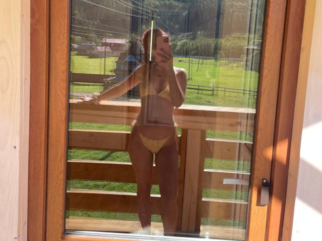 AlexXas in a yellow bikini, standing in front of a glass door, reflection shows her tits and pussy, holding phone, wooden balcony, sunny day, green grass outside.