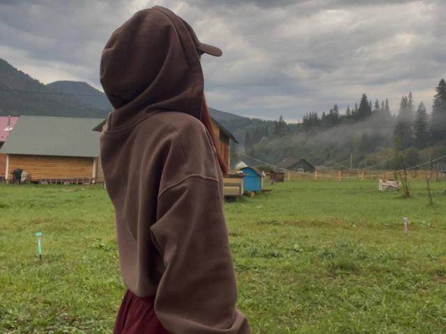 AlexXas, un sweat à capuche marron avec une casquette, dos tourné, montrant ses fesses dans un pantalon rouge, debout dans un champ herbeux, ciel nuageux, maison en bois, et forêt brumeuse en arrière-plan..