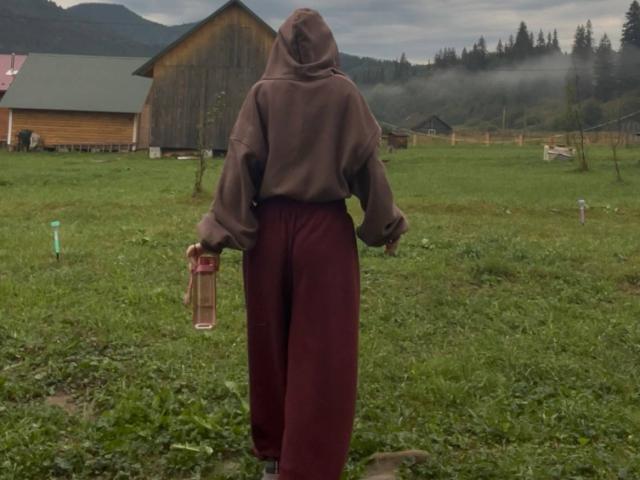 AlexXas with a brown hoodie and maroon pants, holding a pink cup, standing in a grassy field, facing away, in a rural, foggy, mountainous area.