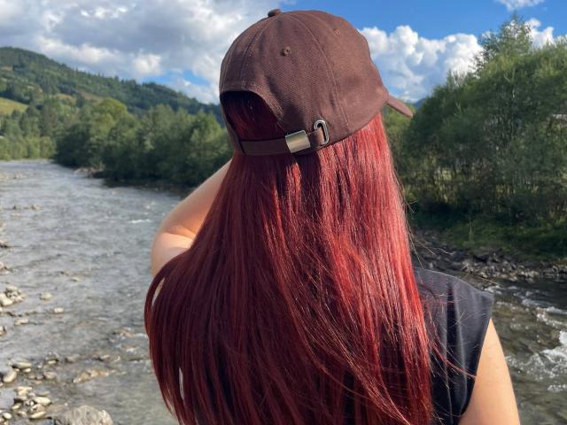 AlexXas, con lunghi capelli rossi, un berretto marrone e una maglietta nera, in piedi vicino a un fiume roccioso, rivolto di spalle, con la luce del sole sui capelli, una foresta verde e un cielo blu con nuvole sullo sfondo..