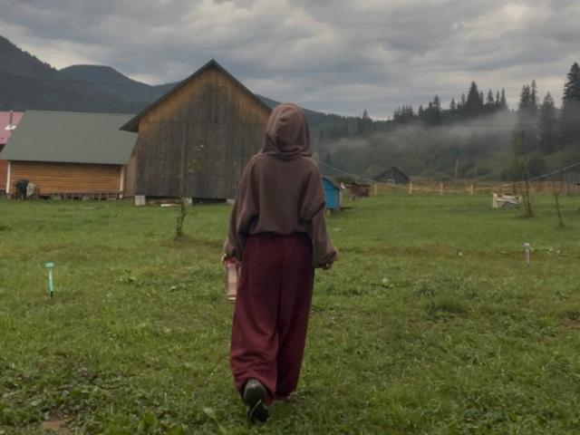 AlexXas with a brown hood and red pants walks away from a wooden barn, holding a small bottle, on a grassy, foggy field with a misty mountain backdrop.
