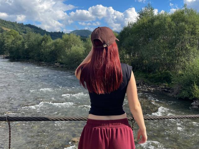 AlexXas, avec des cheveux roux, un haut noir, un pantalon bordeaux et une casquette marron, se tient au bord d'une rivière, dos à la caméra, le bras droit reposant sur une corde rouillée. Une rivière et des arbres sont visibles en arrière-plan..
