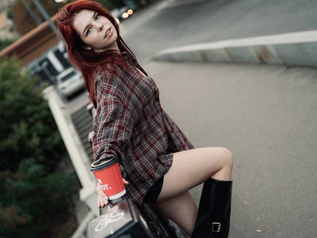 AlexXas with red hair, wearing a plaid shirt, black shorts, and black boots, holding a red cup, sitting on a car, showing her bare legs and a sexy smile.