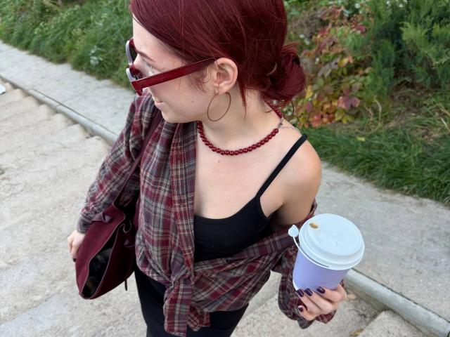 AlexXas with red hair, glasses, hoop earrings, red bead necklace, plaid shirt, black bra, holding coffee cup, maroon bag, walking on concrete path, grassy area.