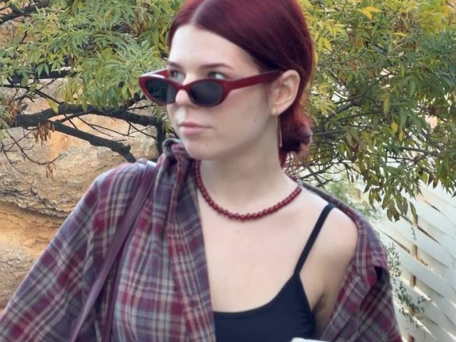 AlexXas, avec des cheveux roux, un débardeur noir, un collier de perles rouges, une chemise à carreaux rouges et des lunettes de soleil rouges, regardant sur le côté avec une expression légèrement agacée..