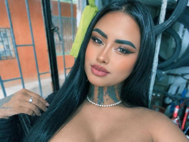LairaHearvy has long black hair, thick eyebrows, and full pink lips. She's wearing a pearl necklace, and has a tattoo on her neck. Boobs visible, hands near face.