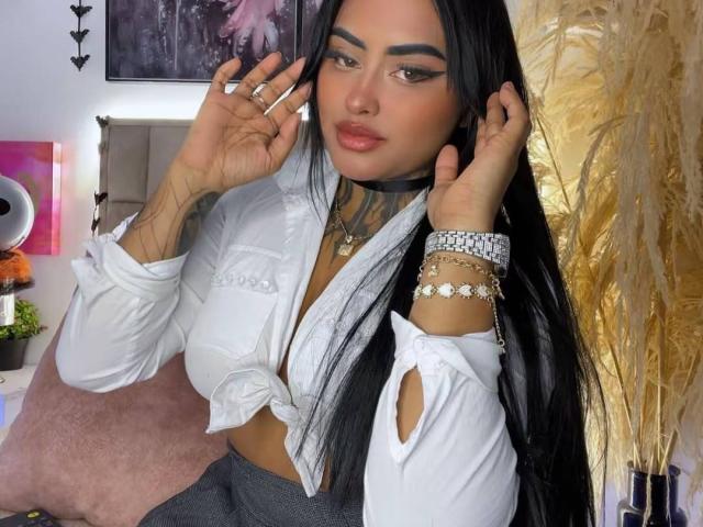 LairaHearvy with long black hair, wearing a white tied-up shirt, showing tattoos, holding her face, and wearing multiple bracelets. Boobs visible through shirt, sitting with a pink pillow.