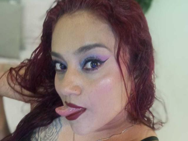 VictoriaBlake with red curly hair, purple eyeliner, and dark lipstick, sticking out her tongue. She has a tattoo on her right shoulder and is looking at the camera.