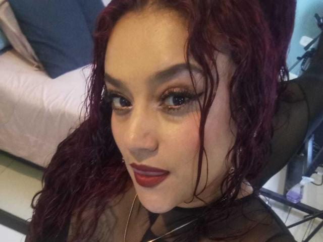 VictoriaBlake with wavy red hair, dark eyeliner, and red lipstick, looking up with a slight smile. She has full breasts and is wearing a black sheer top.