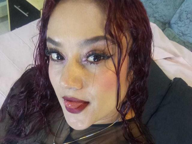 VictoriaBlake has wet red hair, dark eyeliner, and purple eyeshadow. She's sticking her tongue out with a shiny white substance on her face and dark lipstick.