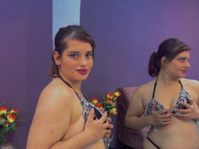 VivianFranco, a chubby woman with brown hair in a ponytail, wearing a black-and-white patterned bikini, holds her tits in front of a mirror, showing her reflection.