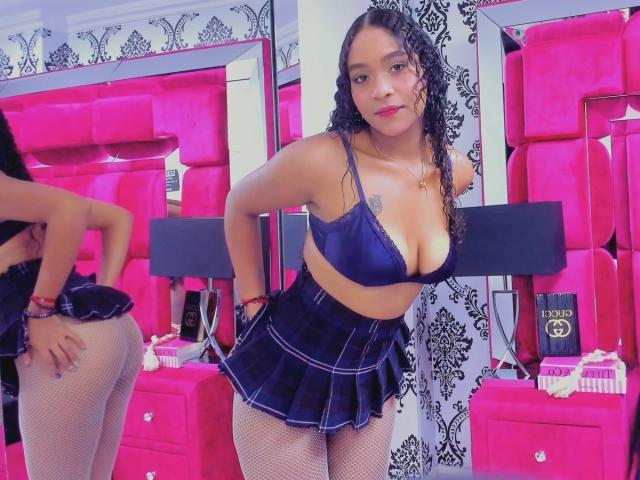 CassidyWinslow with curly black hair, wearing a blue bra, plaid skirt, and fishnet stockings, showing off her tits and ass, standing in a pink, mirrored room.