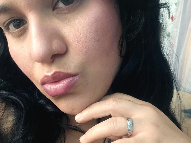 VivianaThomson's face, with pinkish lips, black wavy hair, and a silver ring on her finger, looking directly at the camera. Her skin has a slightly textured, pinkish tone.