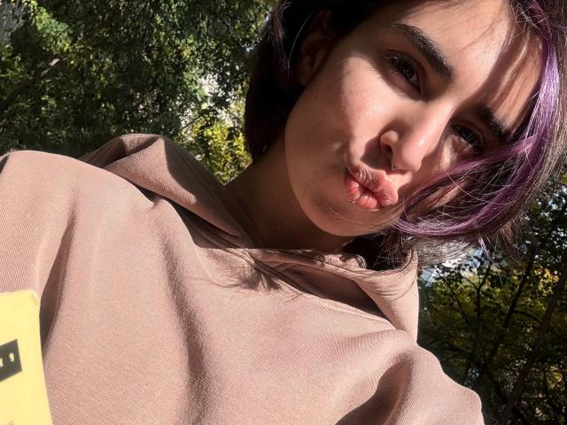 TameikaMacclellan with pouty lips, light brown hoodie, purple hair, and sunlit face, holding a yellow phone in a forest. Her tits are not visible.