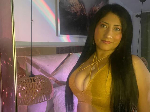 IsabellaBigBoobs, avec de longs cheveux noirs, un soutien-gorge en dentelle jaune, montrant de grandes seins, souriante, dans une pièce avec un éclairage rose et violet, avec une lampe en osier suspendue..