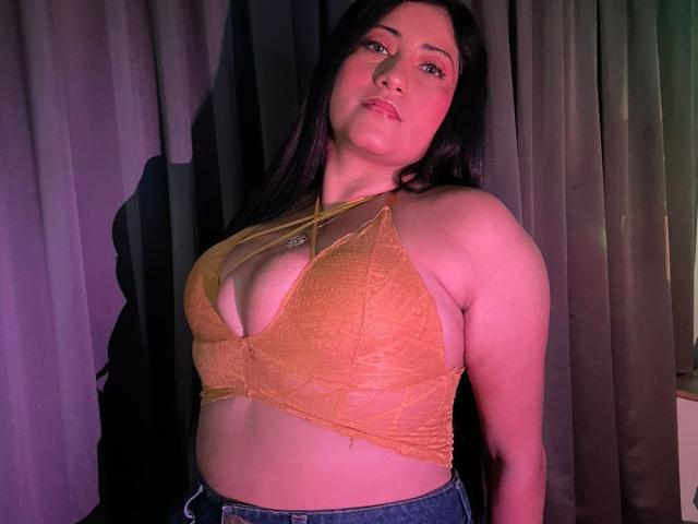 IsabellaBigBoobs with long black hair, orange lace bralette showing her big tits, and blue jeans, looking down with a slight smirk, pinkish-purple light on her face.