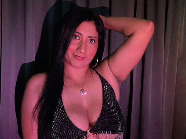 IsabellaBigBoobs mit schwarzen Haaren, trägt ein glitzerndes schwarzes Halstopp, zeigt ihre großen Brüste, Arm erhoben, lächelnd, rosafarbenes Licht, Reifenohrringe, Halskette, sitzt auf einem Stuhl..