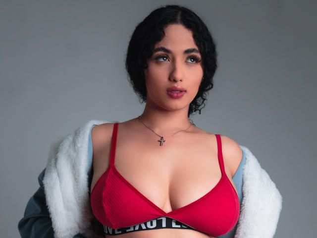 AuroraFerretti with curly black hair, wearing a red bra, showing her big tits, a white fur jacket off her shoulders, and a cross necklace, looking at the camera.