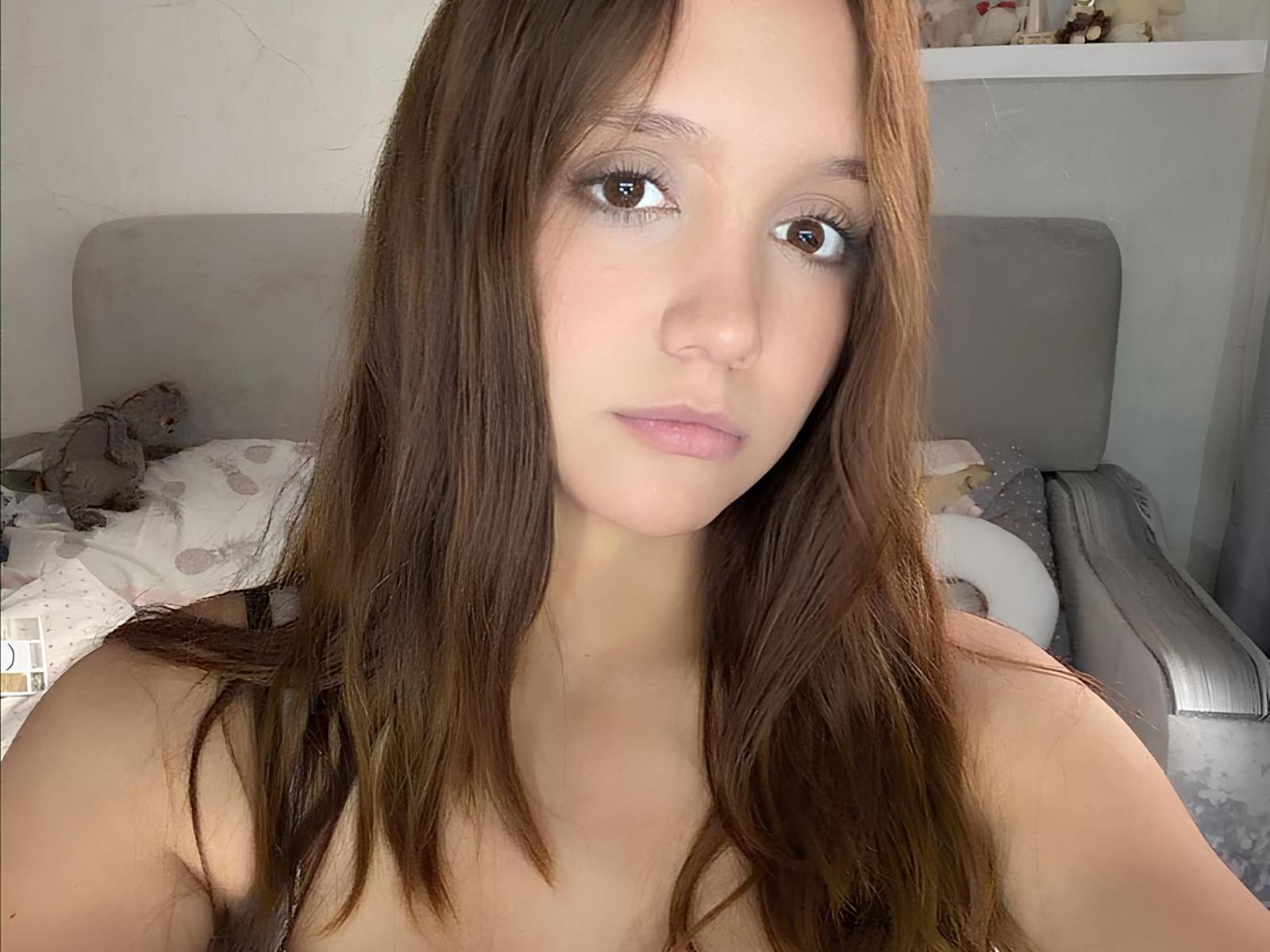 NuPastiva met lang bruin haar, blauwe oogschaduw, roze lippen en lichte huid, kijkt naar de camera, ongetooid, met kleine borsten, in een zacht verlichte slaapkamer..
