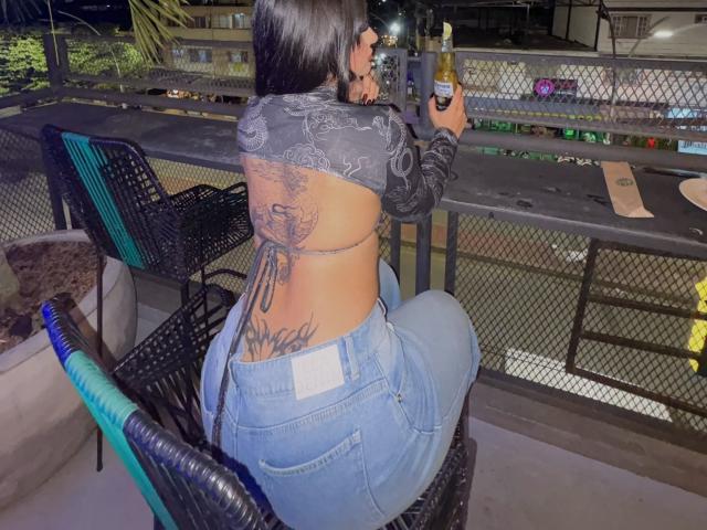 ScarletFeline with tattoos on her back, wearing a black lace top, blue jeans, and holding a drink, sitting at a bar table, her ass facing the camera.