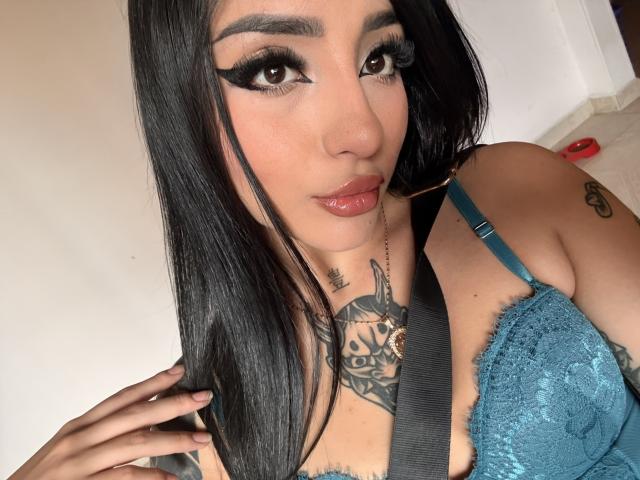ScarletFeline tiene el pelo largo y negro, delineador de ojos grueso y labios rojos brillantes. Lleva un sujetador de encaje color turquesa, mostrando tatuajes en el pecho y el cuello, y tiene una mano cerca de su rostro..
