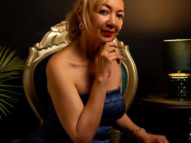 AmelieLux with blonde hair, wearing a strapless blue dress, sitting on an ornate chair, touching her chin, smiling, with red lipstick and hoop earrings.
