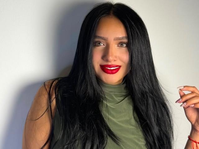 LilyConorr, con lunghi capelli neri, rossetto rosso, top verde senza maniche, che sorride, mano destra con unghie curate, pelle chiara, occhi marroni, braccialetto rosso, su sfondo bianco..
