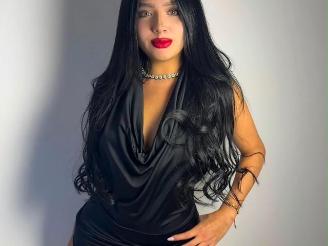 LilyConorr has long black hair, red lipstick, black cowl-neck dress, choker with diamonds, and a bracelet, standing with hands on hips, looking confident.