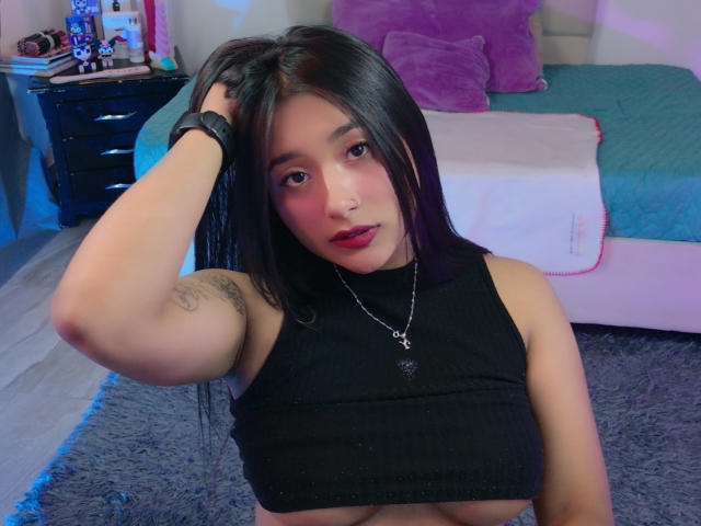 HannahJohnes, avec de longs cheveux noirs, portant un crop top noir qui expose ses seins, une main dans les cheveux, un tatouage sur le bras, du rouge à lèvres, un collier et une montre, assise sur un tapis gris..
