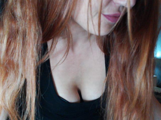 LilouLove has red, wavy hair covering her face, wearing a black top that reveals her cleavage and small tits. Her pink lips are slightly visible.