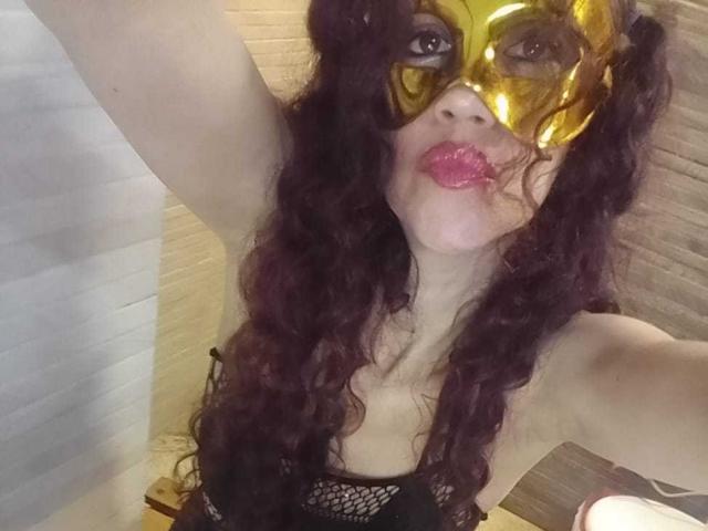 BrendaCathy, con i capelli scuri e ricci, indossa una maschera dorata lucida e rossetto rosa, una maglia nera a rete e alza un braccio per un selfie, mostrando il seno e un'espressione carina e giocosa..