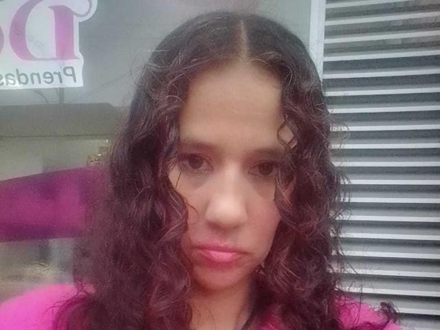BrendaCathy a les cheveux bruns bouclés, du rouge à lèvres rose et une expression sérieuse. Elle porte un haut rose, et ses seins sont légèrement visibles. Ses yeux sont fixés sur l'appareil photo..