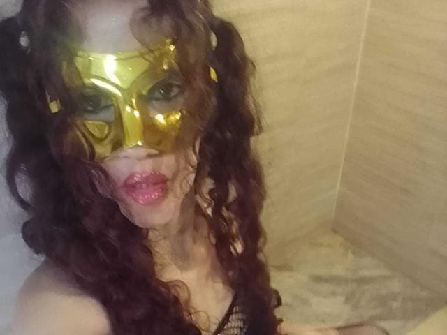 BrendaCathy, met krullend bruin haar, draagt een glanzend geel masker en roze lippenstift, en laat haar borsten zien in een badkamer. Ze zit in een bad met muren betegeld in beige tinten..